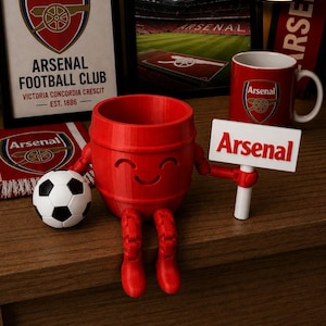 May include: A collection of Arsenal Football Club merchandise, including a framed print, a mug, a scarf, and a 3D-printed red barrel figure with boots and a sign that says "Arsenal". A soccer ball is also present.