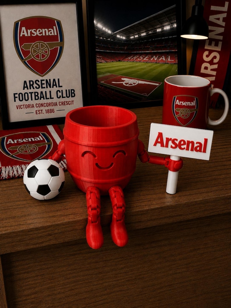May include: A collection of Arsenal Football Club merchandise, including a framed print, a mug, a scarf, and a 3D-printed red barrel figure with boots and a sign that says "Arsenal". A soccer ball is also present.