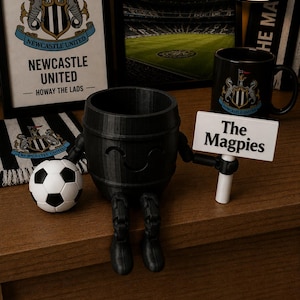 May include: A black barrel-shaped figurine with arms and legs, holding a sign that reads "The Magpies." A soccer ball and a mug with the Newcastle United logo are also present. Additional items include a framed picture and a scarf with the team's logo.