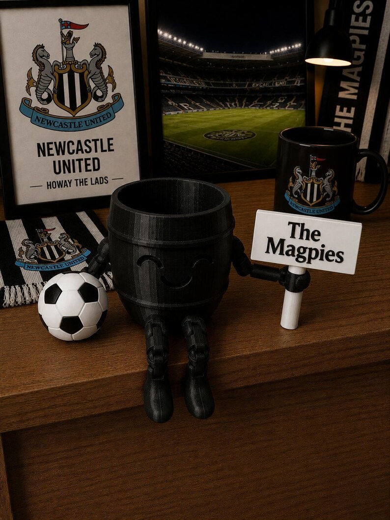 May include: A black barrel-shaped figurine with arms and legs, holding a sign that reads "The Magpies." A soccer ball and a mug with the Newcastle United logo are also present. Additional items include a framed picture and a scarf with the team's logo.