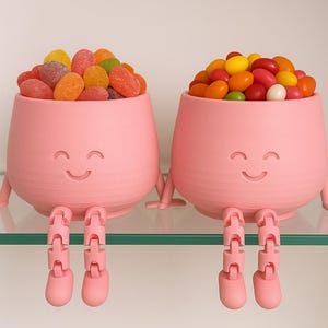 May include: Two pink, smiling, anthropomorphic candy bowls filled with colorful candies. The bowls have legs and arms, and are sitting on a glass shelf. One bowl is filled with gummy candies, the other with jelly beans.
