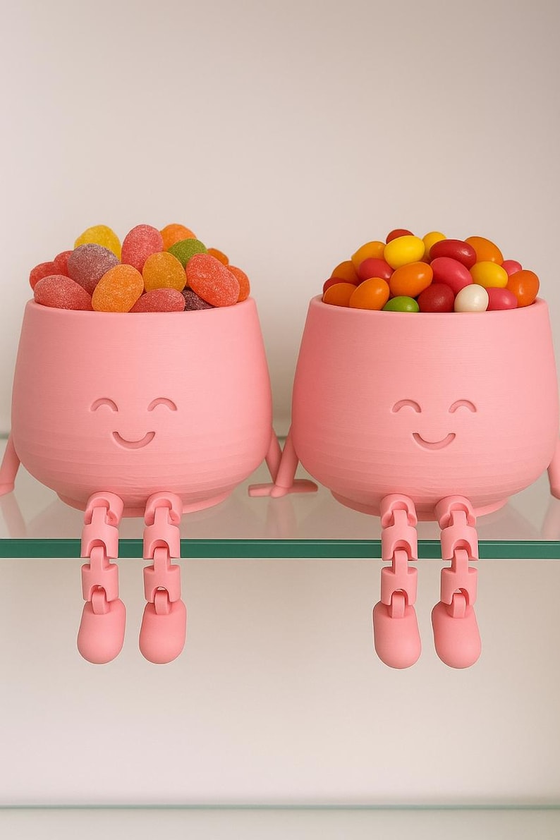 May include: Two pink, smiling, anthropomorphic candy bowls filled with colorful candies. The bowls have legs and arms, and are sitting on a glass shelf. One bowl is filled with gummy candies, the other with jelly beans.