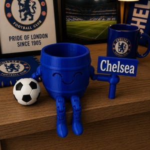 May include: A blue 3D-printed barrel-shaped figure with a smiling face, holding a sign that says "Chelsea." A soccer ball and Chelsea Football Club memorabilia are nearby. The figure has articulated legs.