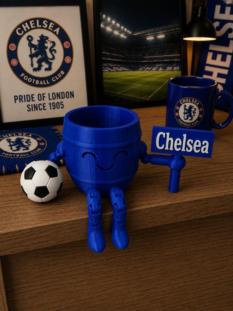 May include: A blue 3D-printed barrel-shaped figure with a smiling face, holding a sign that says "Chelsea." A soccer ball and Chelsea Football Club memorabilia are nearby. The figure has articulated legs.