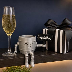 Puede incluir: Una copa de champ&aacute;n con l&iacute;quido burbujeante est&aacute; junto a una figura gris en forma de barril con brazos y piernas, que sostiene un letrero que dice "Felicidades". Tambi&eacute;n hay una caja de regalo a rayas blancas y negras con un lazo negro.
