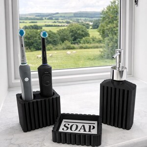 May include: A black bathroom set featuring a soap dish with "SOAP" text, a soap dispenser, and a toothbrush holder with two electric toothbrushes. The set is placed on a white countertop near a window with a scenic view.