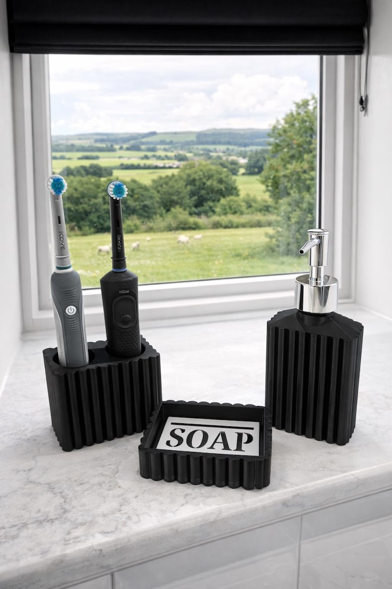 May include: A black bathroom set featuring a soap dish with "SOAP" text, a soap dispenser, and a toothbrush holder with two electric toothbrushes. The set is placed on a white countertop near a window with a scenic view.