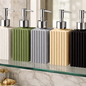 May include: Five rectangular soap dispensers in white, green, gray, beige, and black. Each dispenser has a ribbed design and a silver pump. The dispensers are arranged on a glass shelf in front of a marble wall.