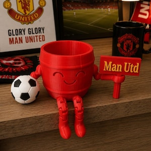 May include: A red 3D-printed barrel-shaped figure with a smiling face, holding a sign that reads "Man Utd." A soccer ball sits next to it. The figure has articulated legs and arms. In the background are Manchester United themed items.