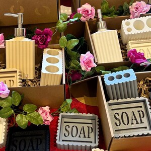 May include: A collection of bathroom accessories, including soap dispensers, toothbrush holders, and soap dishes, presented in cardboard boxes. The items are in neutral tones, with a ribbed design. The word "SOAP" is visible on the soap dishes. Pink roses and greenery add a decorative touch.