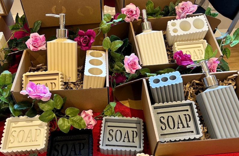May include: A collection of bathroom accessories, including soap dispensers, toothbrush holders, and soap dishes, presented in cardboard boxes. The items are in neutral tones, with a ribbed design. The word "SOAP" is visible on the soap dishes. Pink roses and greenery add a decorative touch.