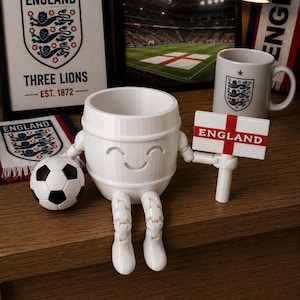 May include: A white, smiling barrel-shaped figurine with articulated legs and arms, holding a sign that reads "ENGLAND." A soccer ball and a mug with the England emblem are also present, along with framed artwork and a scarf.