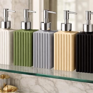 May include: Five rectangular soap dispensers in white, green, gray, beige, and black. Each dispenser has a ribbed design and a silver pump. The dispensers are arranged on a glass shelf in front of a marble wall.
