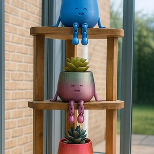 May include: Three smiling, colorful planters with attached legs and arms, each holding a succulent plant. The planters are blue, rainbow-colored, and orange, and are arranged on a wooden shelf unit. The background includes a brick wall and a window.
