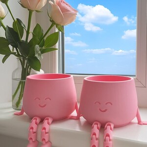 May include: Two pink, smiling face planters with attached legs sit on a windowsill. A vase of pink roses is to the left, and a bright blue sky with clouds is visible through the window.
