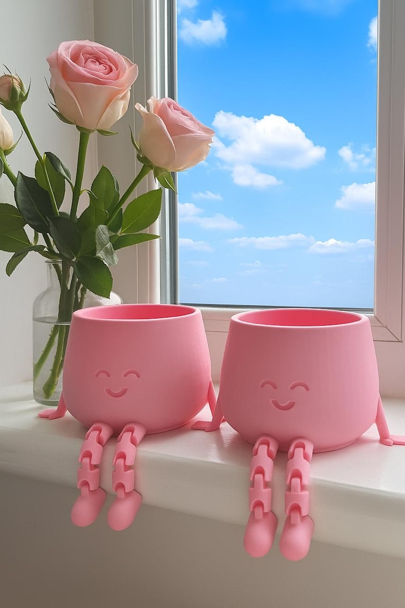 May include: Two pink, smiling face planters with attached legs sit on a windowsill. A vase of pink roses is to the left, and a bright blue sky with clouds is visible through the window.