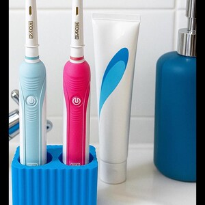 Puede incluir: Dos cepillos de dientes el&eacute;ctricos, uno azul y otro rosa, con la marca "KOYE" en el mango, en un soporte azul. Un tubo de pasta de dientes y un dispensador azul tambi&eacute;n son visibles.