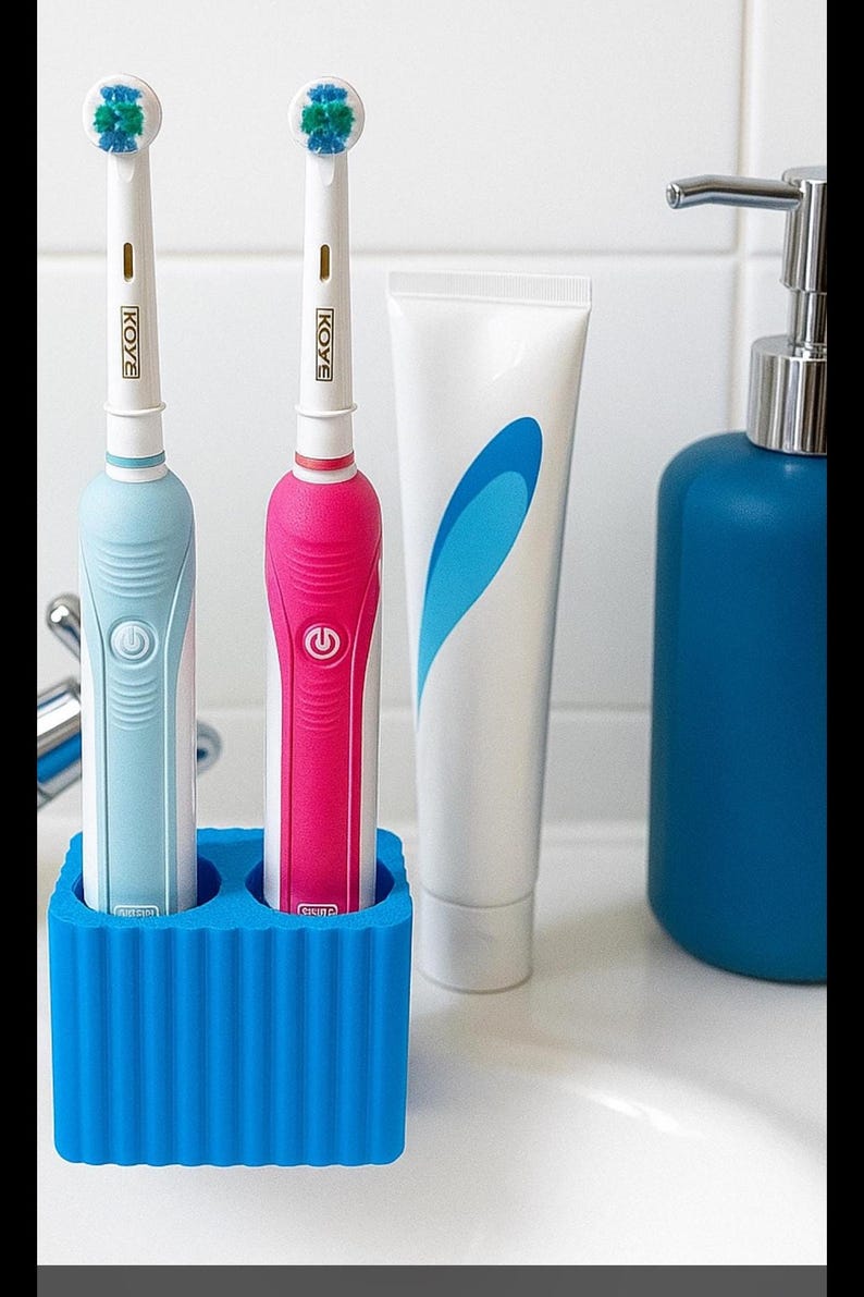 Puede incluir: Dos cepillos de dientes el&eacute;ctricos, uno azul y otro rosa, con la marca "KOYE" en el mango, en un soporte azul. Un tubo de pasta de dientes y un dispensador azul tambi&eacute;n son visibles.
