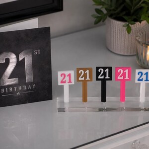 Puede incluir: Una tarjeta de cumplea&ntilde;os n&uacute;mero 21 en negro y plateado se muestra junto a un conjunto de adornos para pasteles con el n&uacute;mero 21. Los adornos son blancos, marrones, negros, rosas y azules. Un soporte acr&iacute;lico transparente los mantiene en posici&oacute;n vertical.