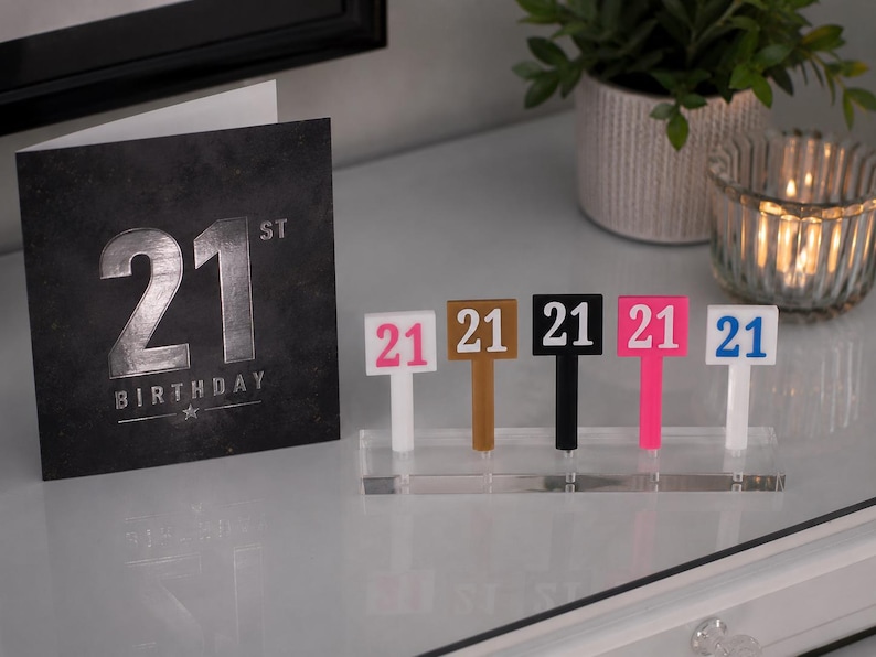 Puede incluir: Una tarjeta de cumplea&ntilde;os n&uacute;mero 21 en negro y plateado se muestra junto a un conjunto de adornos para pasteles con el n&uacute;mero 21. Los adornos son blancos, marrones, negros, rosas y azules. Un soporte acr&iacute;lico transparente los mantiene en posici&oacute;n vertical.
