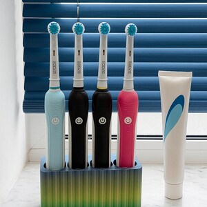 Puede incluir: Cuatro cepillos de dientes el&eacute;ctricos en un soporte, cada uno con un mango de diferente color: azul claro, negro, marr&oacute;n oscuro y rosa. El soporte es un degradado de verde, amarillo y azul. Un tubo de pasta de dientes est&aacute; al lado del soporte.