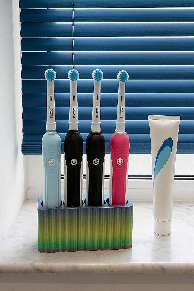 Puede incluir: Cuatro cepillos de dientes el&eacute;ctricos en un soporte, cada uno con un mango de diferente color: azul claro, negro, marr&oacute;n oscuro y rosa. El soporte es un degradado de verde, amarillo y azul. Un tubo de pasta de dientes est&aacute; al lado del soporte.