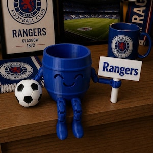 May include: A blue 3D-printed barrel-shaped figure with a smiling face, holding a sign that reads "Rangers." A soccer ball and a mug with the Rangers logo are also visible. The image includes framed artwork and a scarf with the Rangers logo.