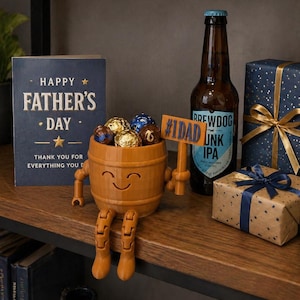 Puede incluir: Una composici&oacute;n para el D&iacute;a del Padre con un recipiente de madera en forma de barril lleno de bombones. Una tarjeta con la frase "Happy Father's Day" y una botella de cerveza est&aacute;n presentes, junto con regalos envueltos.