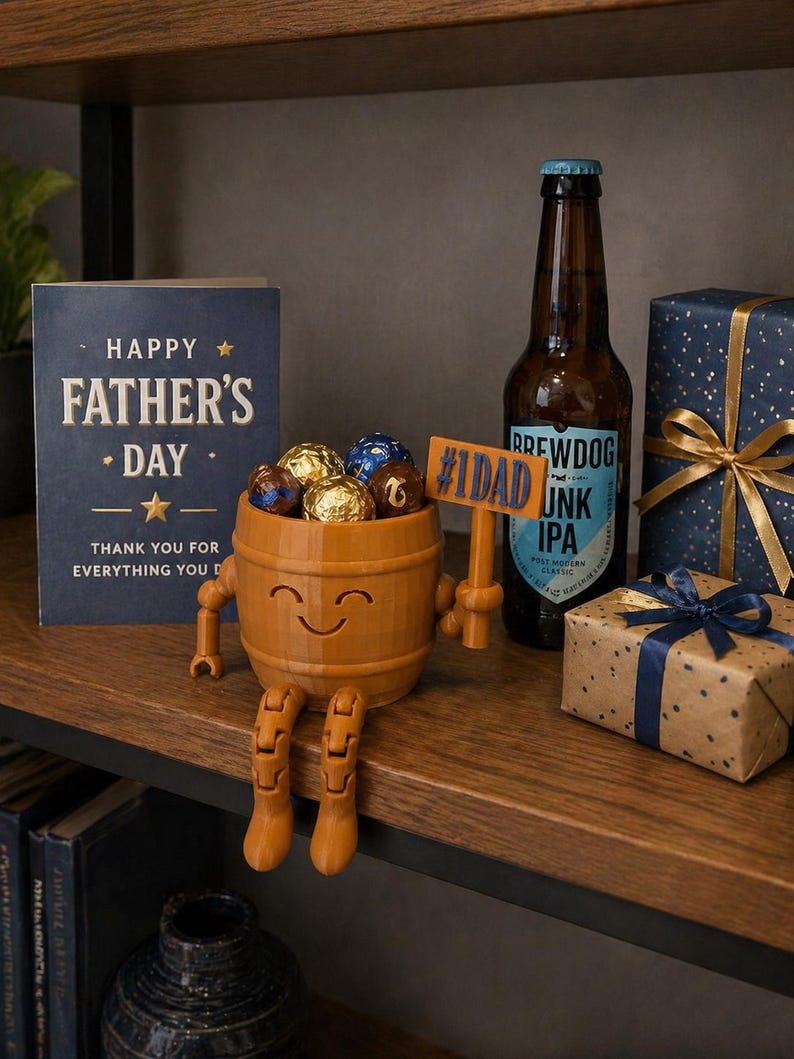 Puede incluir: Una composici&oacute;n para el D&iacute;a del Padre con un recipiente de madera en forma de barril lleno de bombones. Una tarjeta con la frase "Happy Father's Day" y una botella de cerveza est&aacute;n presentes, junto con regalos envueltos.