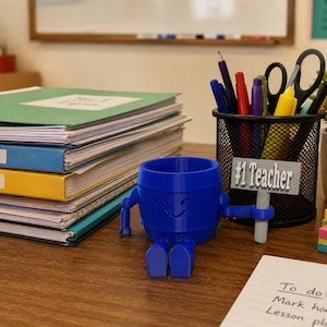 May include: A blue 3D-printed barrel-shaped character figurine sitting on a desk, holding a gray sign that reads #1 Teacher. The figure is positioned next to a black mesh pencil holder filled with pens, pencils, and scissors, and a stack of notebooks.