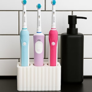 Puede incluir: Una composici&oacute;n de ba&ntilde;o con tres cepillos de dientes el&eacute;ctricos azules, morados y rosas, en un soporte blanco. Un dispensador de jab&oacute;n negro a la derecha. Fondo de azulejos blancos.