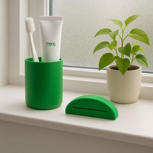 May include: A bright green toothbrush holder with a white toothbrush and a tube of toothpaste. A green toothpaste squeezer sits in front of the holder. A small potted plant is in the background. The toothpaste tube has the text "ne%" on it.