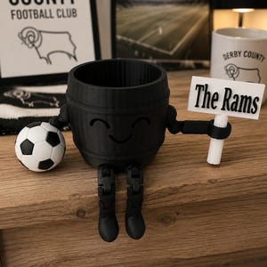 May include: A black 3D-printed planter shaped like a barrel with arms, legs, and a smiling face. It holds a soccer ball and a sign that reads "The Rams." The planter is on a wooden surface, with Derby County Football Club items in the background.