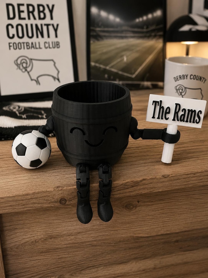 May include: A black 3D-printed planter shaped like a barrel with arms, legs, and a smiling face. It holds a soccer ball and a sign that reads "The Rams." The planter is on a wooden surface, with Derby County Football Club items in the background.