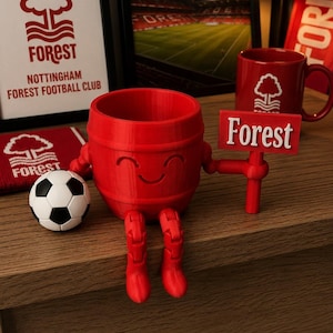 May include: A red, barrel-shaped figurine with a smiling face, holding a sign that reads "Forest." A soccer ball and a red mug with a tree logo are also present. The image includes framed artwork and a banner, all featuring the Nottingham Forest Football Club logo.