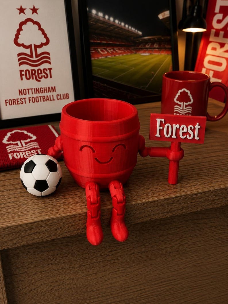 May include: A red, barrel-shaped figurine with a smiling face, holding a sign that reads "Forest." A soccer ball and a red mug with a tree logo are also present. The image includes framed artwork and a banner, all featuring the Nottingham Forest Football Club logo.