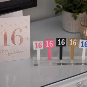 Puede incluir: Una tarjeta de cumplea&ntilde;os con el n&uacute;mero 16 en oro rosa y las palabras "Happy Birthday". Junto a ella, cinco letreros coloridos con el n&uacute;mero 16 en rosa, negro, dorado y azul. Los letreros est&aacute;n sobre un soporte acr&iacute;lico transparente.
