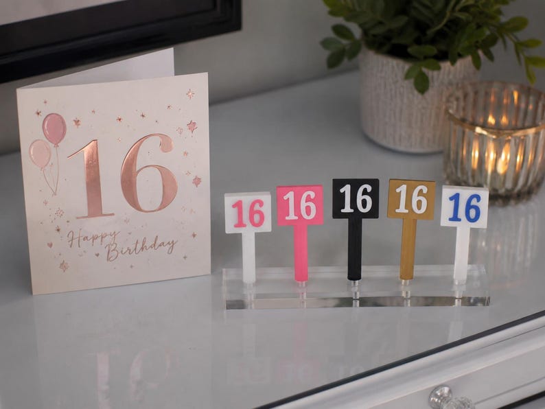Puede incluir: Una tarjeta de cumplea&ntilde;os con el n&uacute;mero 16 en oro rosa y las palabras "Happy Birthday". Junto a ella, cinco letreros coloridos con el n&uacute;mero 16 en rosa, negro, dorado y azul. Los letreros est&aacute;n sobre un soporte acr&iacute;lico transparente.