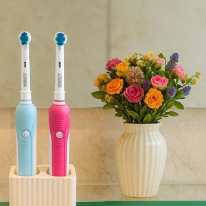 Puede incluir: Dos cepillos de dientes el&eacute;ctricos, uno azul claro y otro rosa, se muestran en un soporte color crema. Los cepillos de dientes tienen mangos blancos y cabezales azules. Un jarr&oacute;n con flores coloridas est&aacute; cerca.
