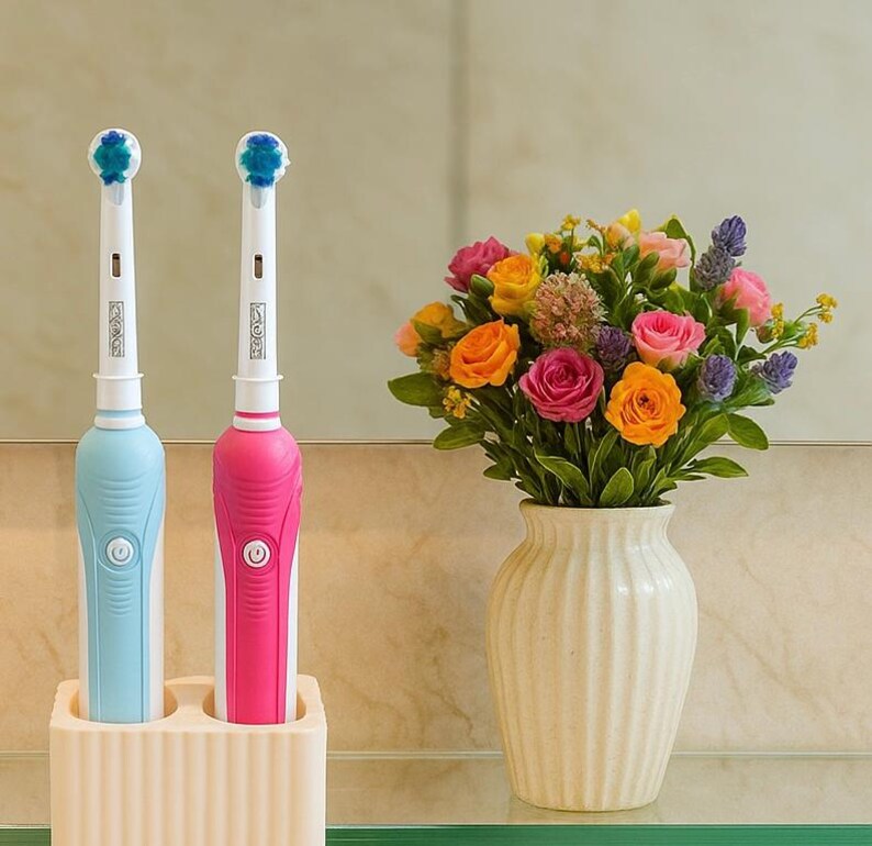 Puede incluir: Dos cepillos de dientes el&eacute;ctricos, uno azul claro y otro rosa, se muestran en un soporte color crema. Los cepillos de dientes tienen mangos blancos y cabezales azules. Un jarr&oacute;n con flores coloridas est&aacute; cerca.