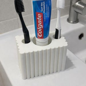 May include: A white toothbrush holder with a scalloped design, holding a tube of Colgate toothpaste, a black toothbrush, and a white toothbrush. The toothpaste tube has blue and red branding.