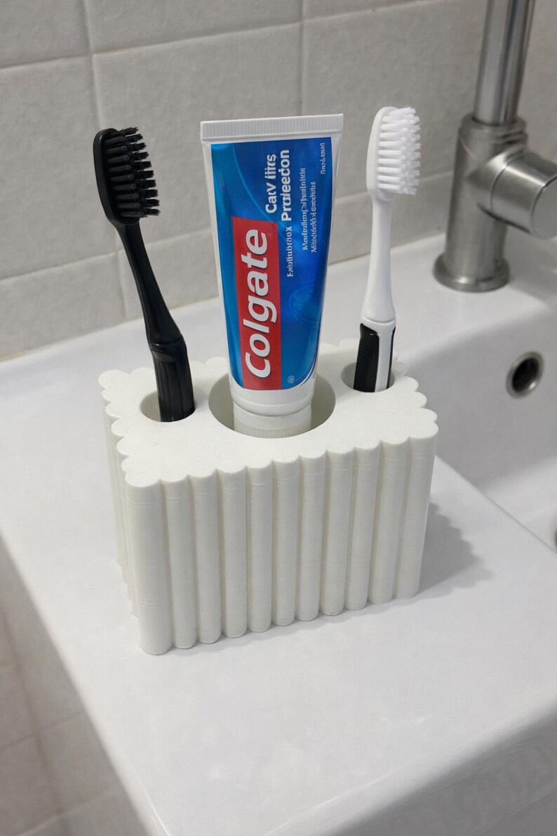 May include: A white toothbrush holder with a scalloped design, holding a tube of Colgate toothpaste, a black toothbrush, and a white toothbrush. The toothpaste tube has blue and red branding.