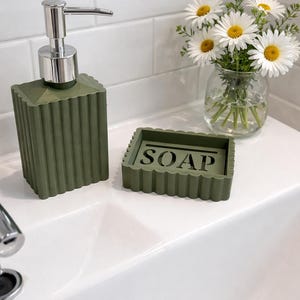 May include: Olive green soap dispenser with a silver pump and matching soap dish. The soap dish has the word "SOAP" carved into it. A small glass vase with white daisies sits nearby.