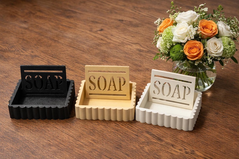 May include: Three decorative soap dishes in black, cream, and white, each with a scalloped edge and a cutout design spelling "SOAP". A bouquet of flowers with orange and white roses sits in the background.