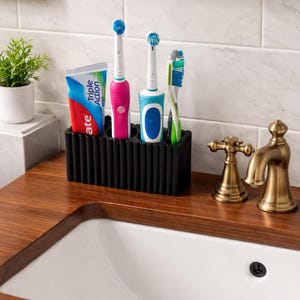 Puede incluir: Un soporte negro para cepillos de dientes con un tubo de pasta de dientes, dos cepillos de dientes el&eacute;ctricos y un cepillo de dientes manual. Los cepillos de dientes son rosas, azules y verdes. El tubo de pasta de dientes es rojo, blanco y azul. El soporte est&aacute; sobre una encimera de madera.