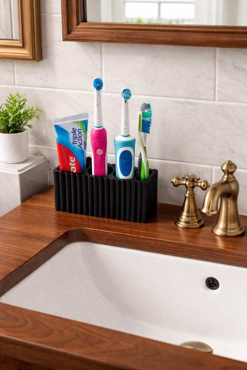 Puede incluir: Un soporte negro para cepillos de dientes con un tubo de pasta de dientes, dos cepillos de dientes el&eacute;ctricos y un cepillo de dientes manual. Los cepillos de dientes son rosas, azules y verdes. El tubo de pasta de dientes es rojo, blanco y azul. El soporte est&aacute; sobre una encimera de madera.