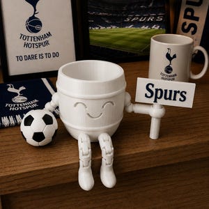 May include: A white 3D-printed Tottenham Hotspur character with a smiling face, holding a sign that says "Spurs." A soccer ball and a mug with the team logo are also visible. The background includes framed pictures and a scarf with the team's logo.