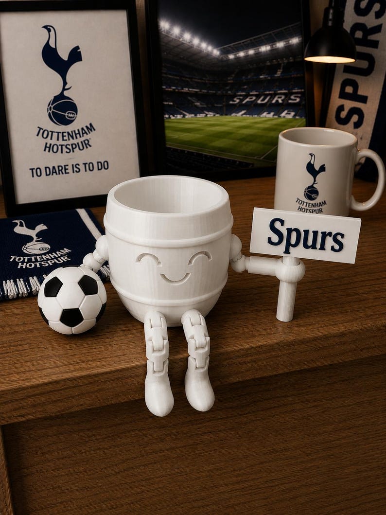 May include: A white 3D-printed Tottenham Hotspur character with a smiling face, holding a sign that says "Spurs." A soccer ball and a mug with the team logo are also visible. The background includes framed pictures and a scarf with the team's logo.