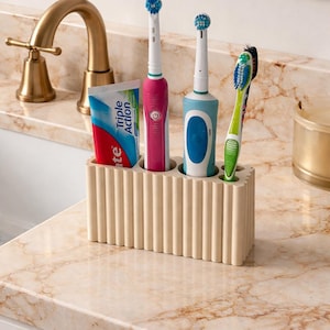 Puede incluir: Un soporte beige y acanalado para cepillos de dientes sobre una encimera de m&aacute;rmol, que contiene un tubo de pasta de dientes, dos cepillos de dientes el&eacute;ctricos y un cepillo de dientes manual. Los cepillos de dientes son rosas, azules y verdes. Un grifo dorado est&aacute; en el fondo.
