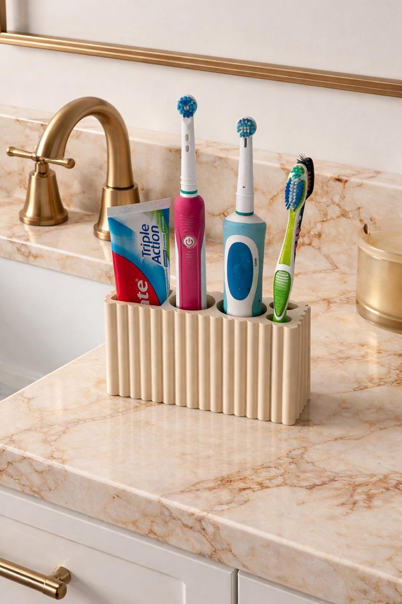 Puede incluir: Un soporte beige y acanalado para cepillos de dientes sobre una encimera de m&aacute;rmol, que contiene un tubo de pasta de dientes, dos cepillos de dientes el&eacute;ctricos y un cepillo de dientes manual. Los cepillos de dientes son rosas, azules y verdes. Un grifo dorado est&aacute; en el fondo.